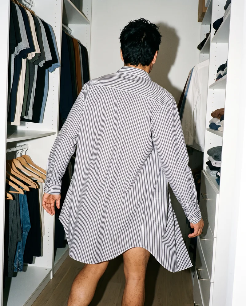 Rear view of a model wearing a striped nightshirt in a walk-in closet, with one leg stepping forward.