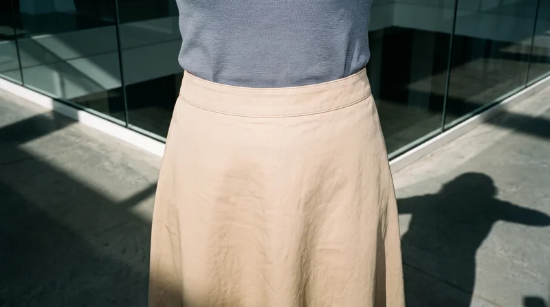 A woman wears a khaki cotton maxi skirt, shown from the front in a waist-level crop focused on the waistline. A skyscraper glass atrium serves as the backdrop.