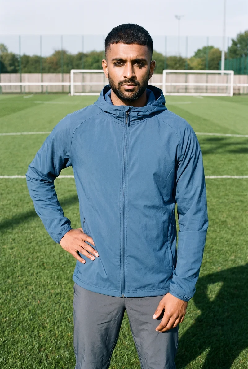 A 20-year-old Indian man is shown full body from the front wearing a blue waterproof ski jacket, with one hand on his hip and a neutral expression. A soccer pitch forms the backdrop.