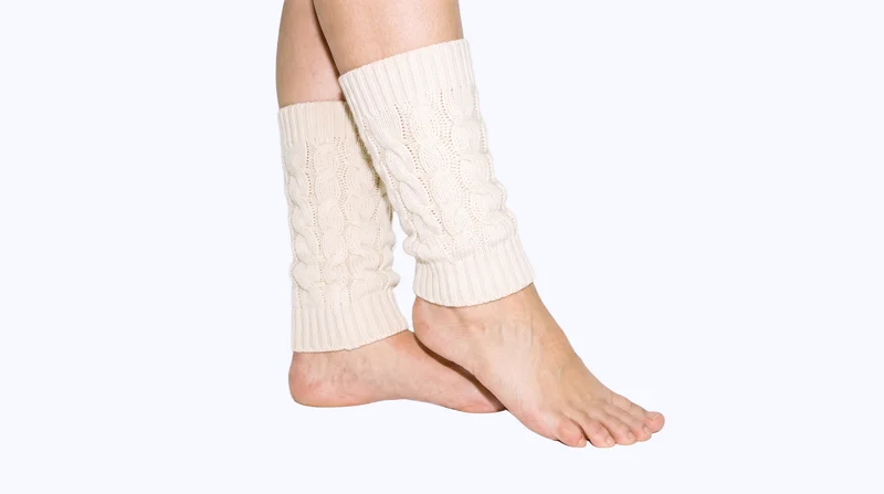 Cream cable-knit leg warmers shown on both feet in a side view, with the ankles crossed. A flat porcelain-colored backdrop surrounds the studio-cutout composition.