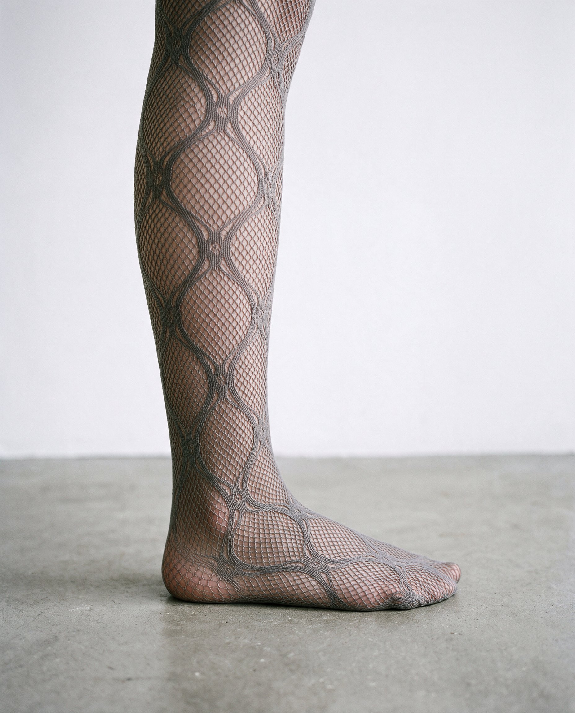 A close-up side view of patterned fishnet tights from the ankle down, with the foot resting flat. The tights are shown against a porcelain studio-wall backdrop.