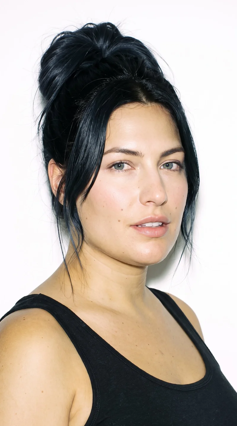 A 34-year-old Caucasian woman with medium olive skin and a cool undertone. Her triangular face has a wide jaw that narrows toward the forehead, emphasizing the lower face. She has thick eyebrows, blue eyes with a cool undertone, and thin lips. Her black hair is gathered into a loose, messy bun with softly undone strands.