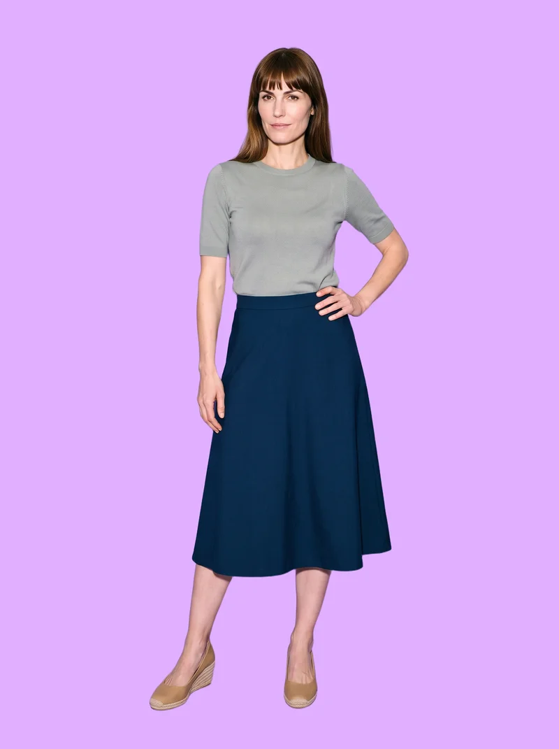 A 43-year-old Caucasian woman is shown full body from the front wearing women's tan wedge espadrilles. She stands with her weight back and a curious expression against a flat mauve backdrop.