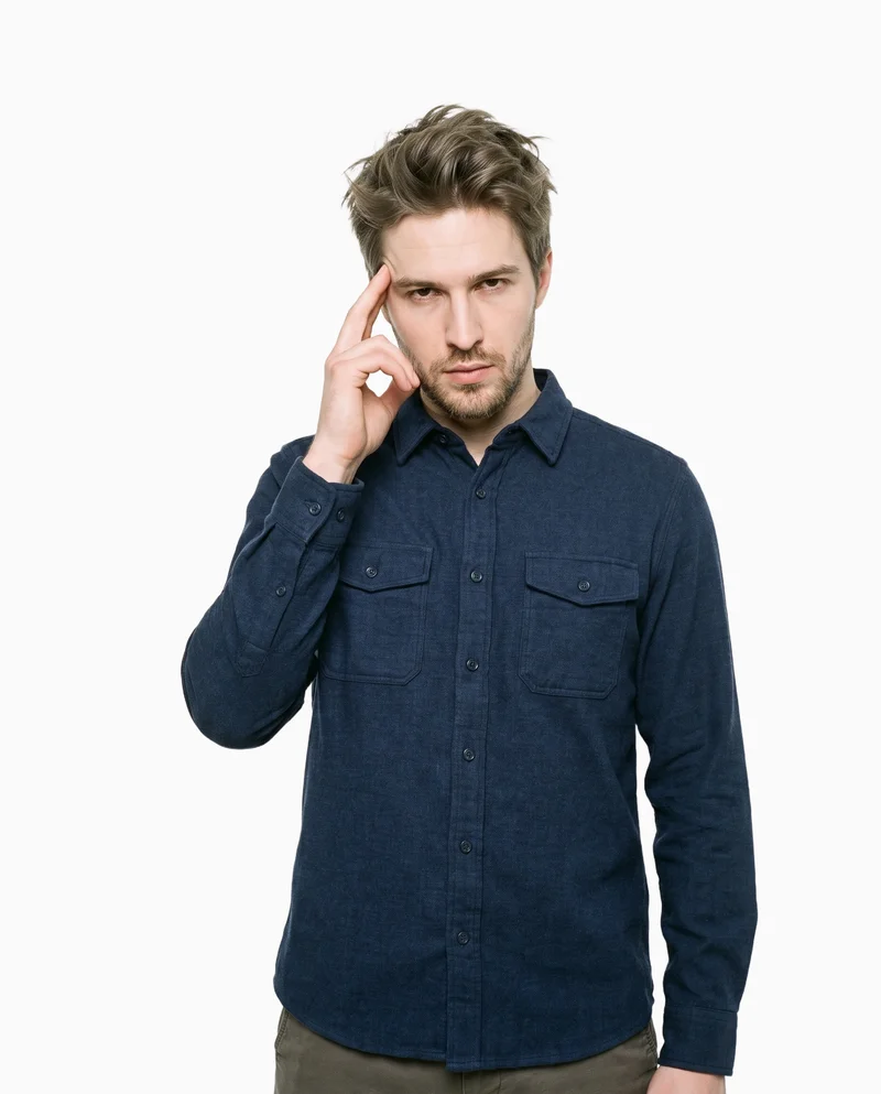 A 25-year-old Caucasian man is shown from the front in an upper-body view wearing a navy flannel overshirt. He holds one hand at his temple with an intense facial expression. A flat Seasalt-colored backdrop fills the background.