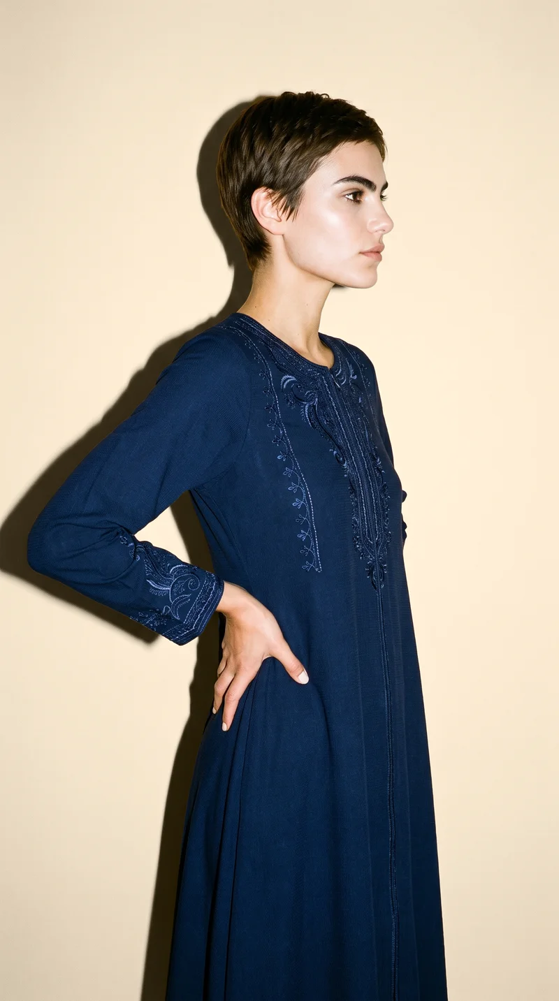 A 25-year-old Caucasian woman wears a navy embroidered abaya, shown in a three-quarter-length side view with her hands on her waist and a neutral expression. A champagne studio-wall backdrop frames the composition.