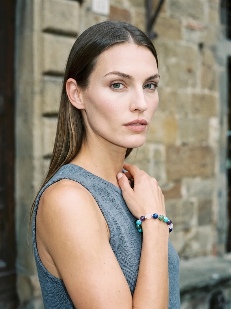 A 25-year-old Caucasian woman is shown from the side in an upper-body composition with one hand resting at her collarbone and a neutral expression. She wears a women's gemstone beaded bracelet. A Tuscany hilltop village serves as the backdrop.