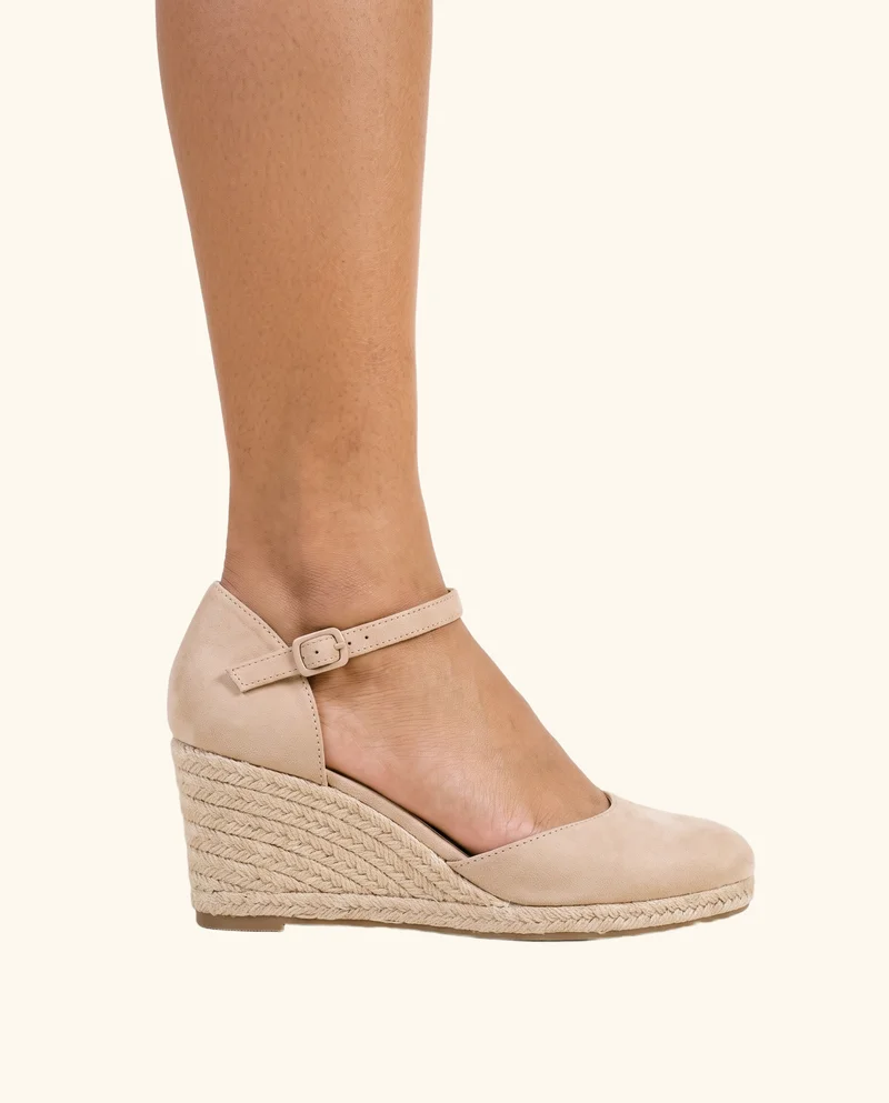 A female ankle wearing nude wedges is shown in side view with the ankle flexed. The footwear is framed from the ankle downward against a creamy white flat-color backdrop.