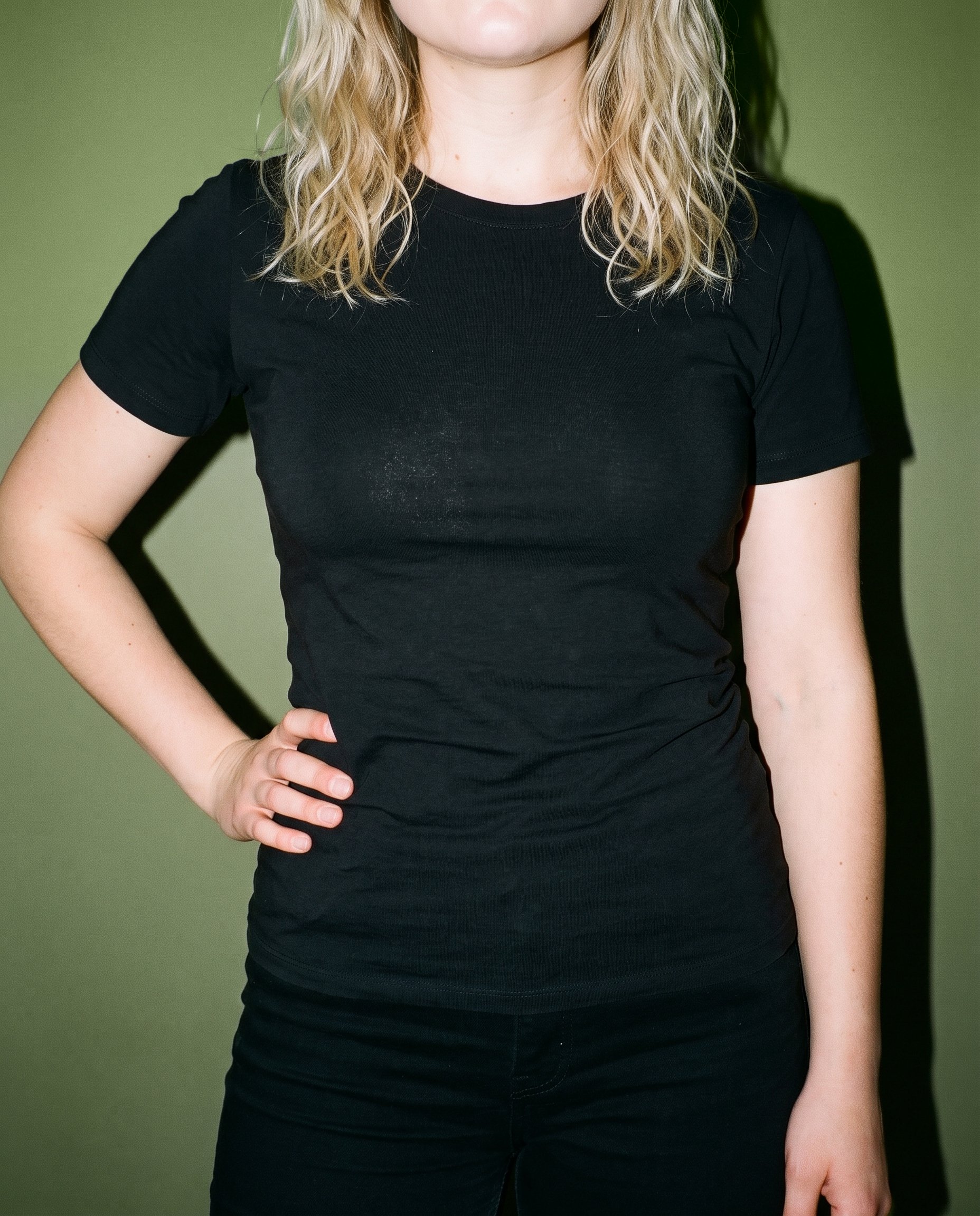 A woman is shown from the torso up wearing a black T-shirt, facing forward with one hand on her hip. An olive grove backdrop is visible behind her.