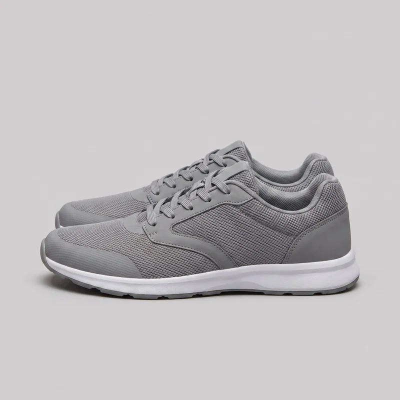 A pair of grey low-top athletic lace-up trainers is shown in a side-oriented three-quarter view. The rounded toes, flat laces, padded collars, mesh-textured upper panels, smooth toe and heel overlays, curved seam lines, and layered cushioned soles are clearly visible.