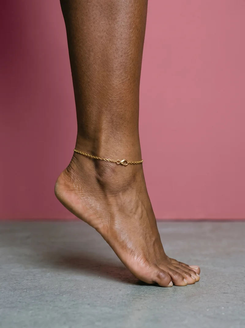 A gold chain anklet worn around an ankle shown from the side as the foot rises onto tiptoe. The image uses a rose-gold studio-wall backdrop.