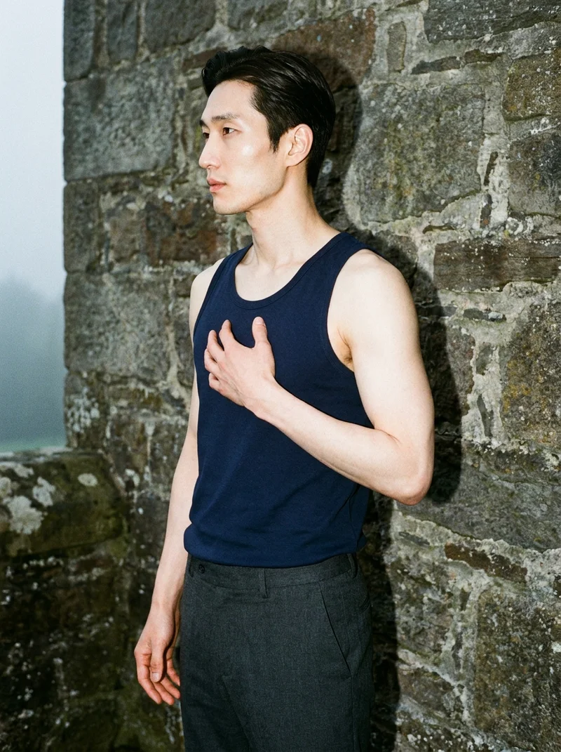 A 34-year-old Korean man is shown from the upper body in side view, wearing a navy blue tank top with his fingers at his chest. The image uses a Scottish castle as the backdrop, and his expression is confident.