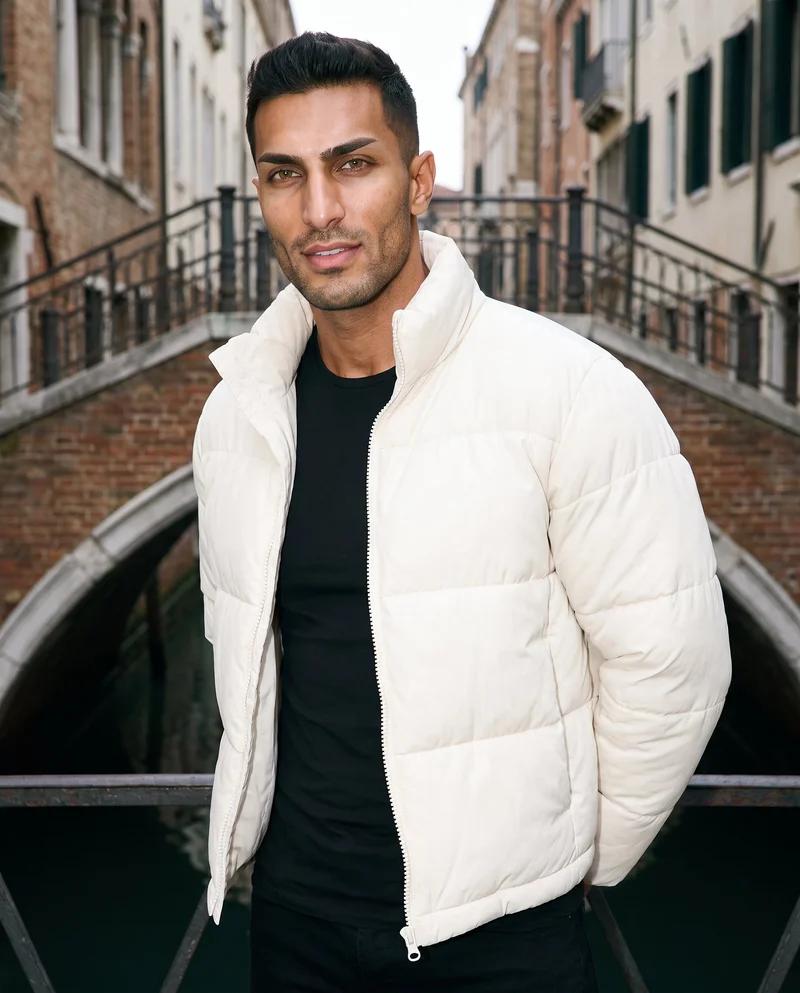 A 31-year-old Arab man wears a men's cream cropped puffer jacket in an upper-body, angled-front view, with his hands behind his back and a smile. A bridge-framed urban view forms the backdrop.