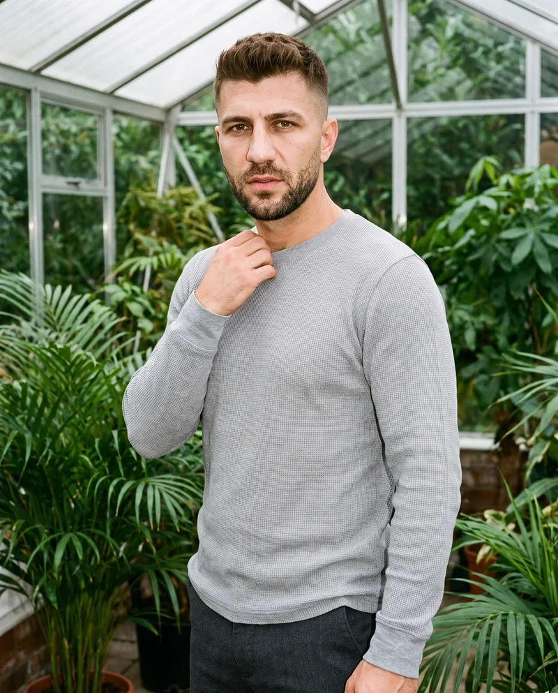 A 37-year-old Turkish man is shown from the upper body at a front three-quarter angle wearing a grey waffle-knit long-sleeve tee. One hand rests at his collarbone, and his expression is neutral. The backdrop is a sunroom.