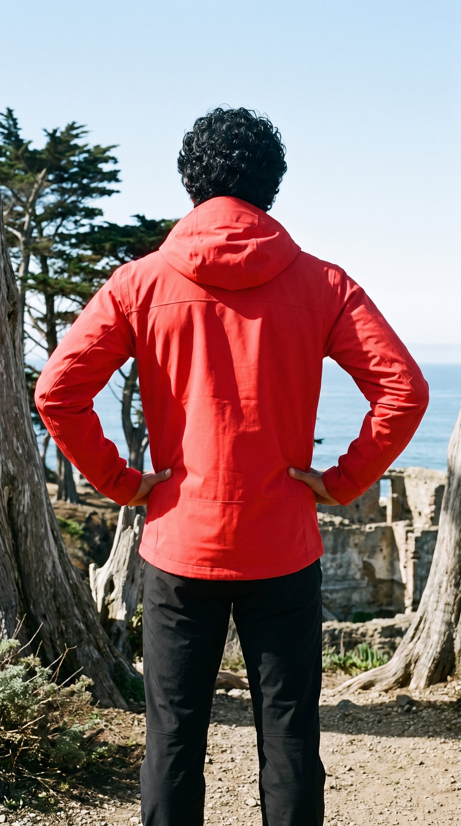 A 26-year-old Southeast Asian man wears a men's red hardshell jacket, shown from the rear in a three-quarter-length view with his hands on his hips. A Coastal Trail Cypress Ruins backdrop is visible behind him.