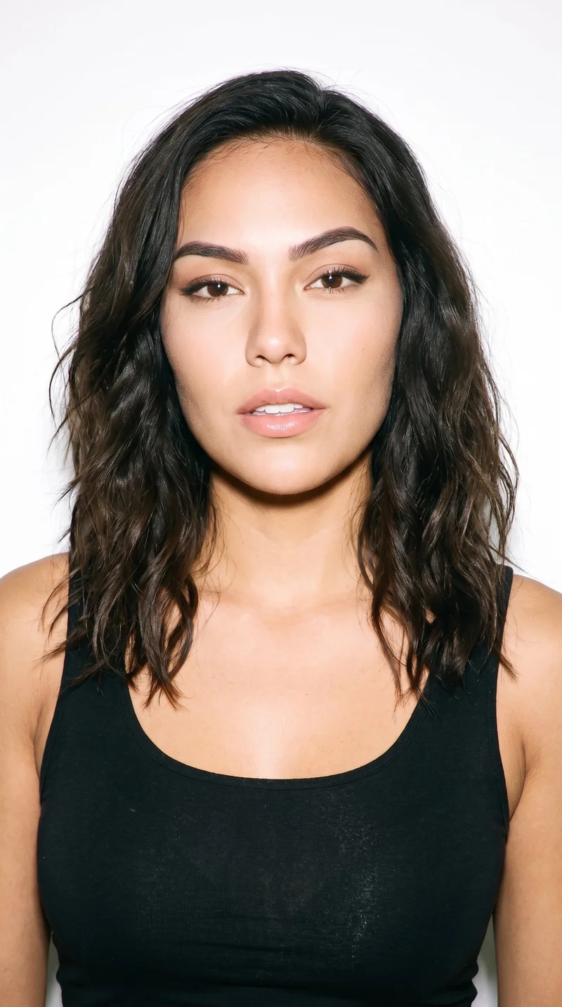 A 24-year-old Indigenous American woman with medium olive skin and a warm greenish-golden undertone. Her heart-shaped face has a wider forehead that narrows to a more pointed chin. She has thick eyebrows, very dark near-black eyes, and medium lips. Her dark-brown hair falls in loose, slightly tousled waves with natural volume.