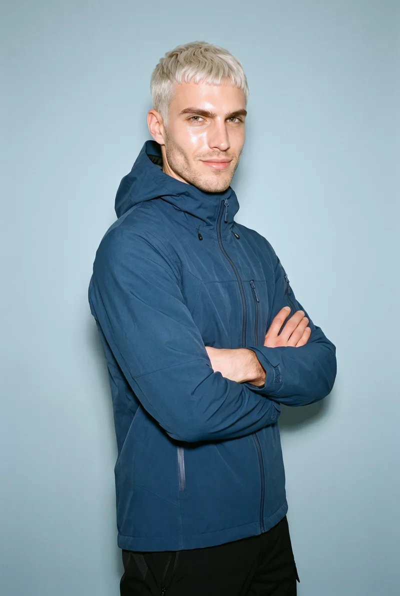 A 29-year-old Caucasian man wears a blue waterproof ski jacket in an upper-body, angled-front view with his arms crossed and a natural expression. A baby blue studio-wall backdrop frames the composition.
