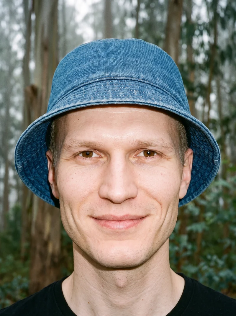 A 38-year-old Slavic man is shown from the head up in a front view, wearing a men's denim bucket hat. He is smiling with his chin raised. An Eucalyptus Fog Park Grove backdrop is visible behind him.