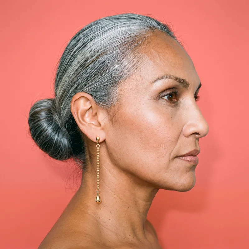 A gold drop earring is shown on the back of an ear in side view. The ear is framed against a coral pink studio-wall backdrop.
