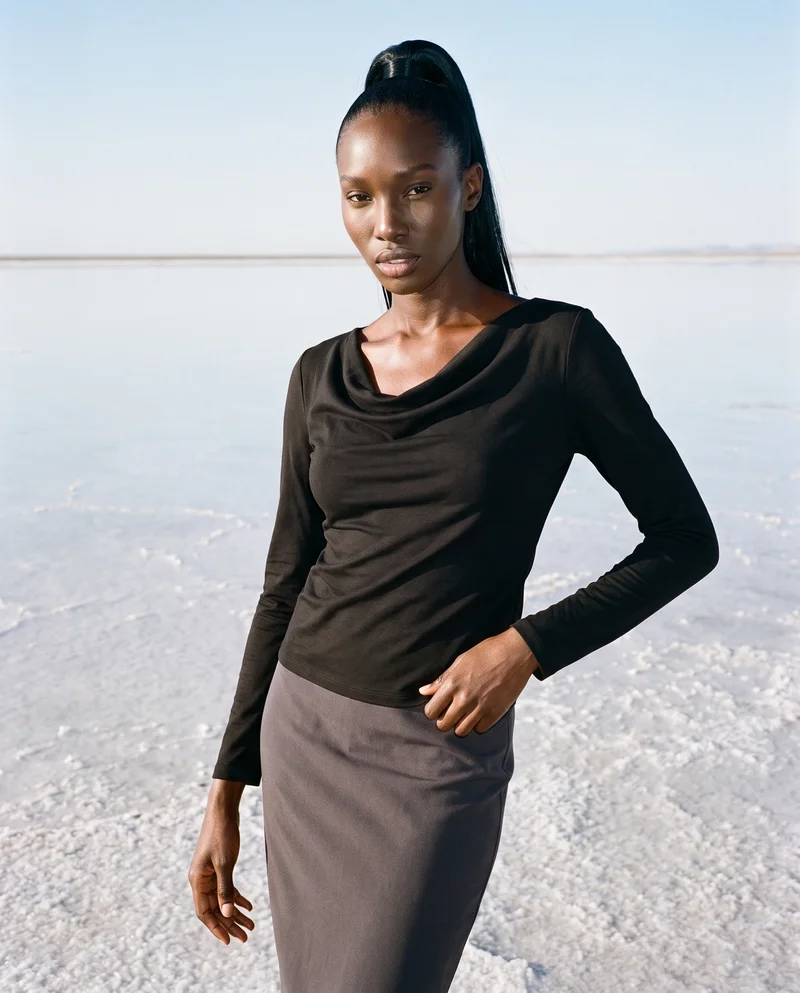 A 29-year-old African woman wears a black cowl-neck top in a front-view, three-quarter-length composition. She stands with her weight shifted back and a soft facial expression, against a salt flats reflection backdrop.