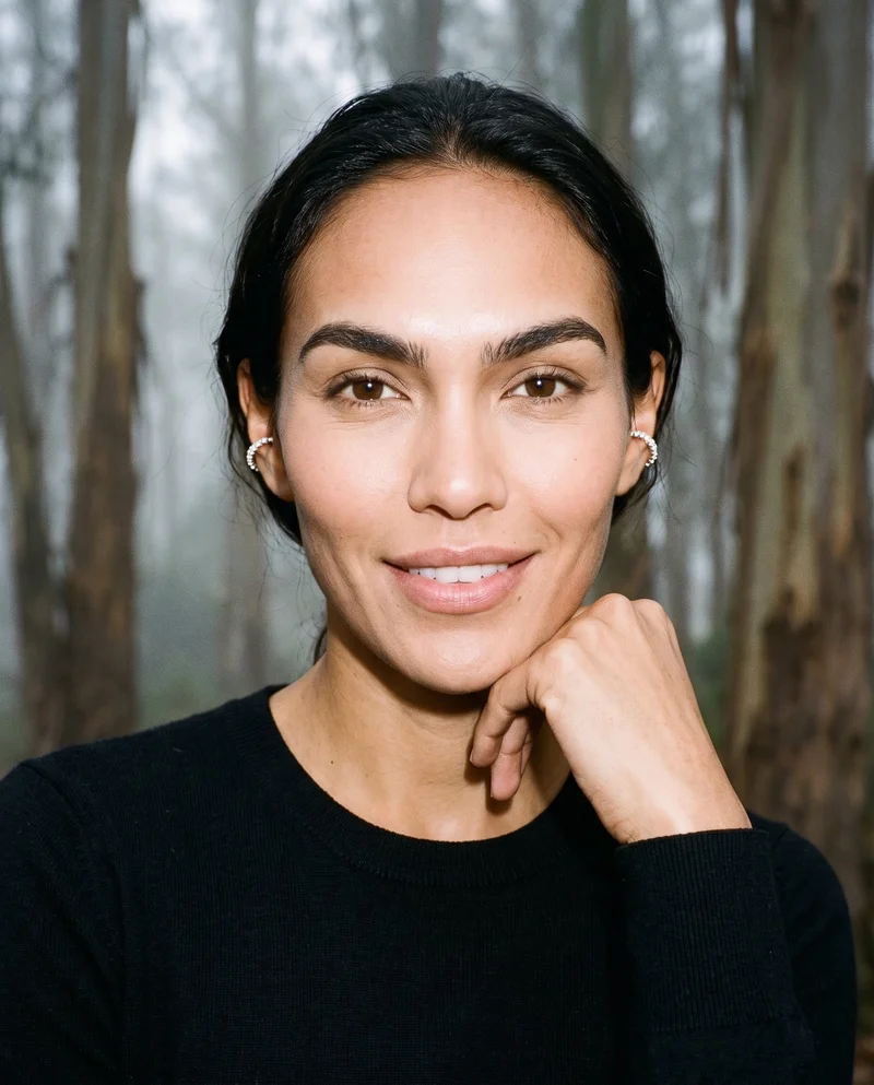 A 36-year-old Indigenous American woman is shown from the bust up in a front-facing view, smiling with her wrist held at her jaw to display crystal ear cuffs. An Eucalyptus Fog Park Grove backdrop appears behind her.