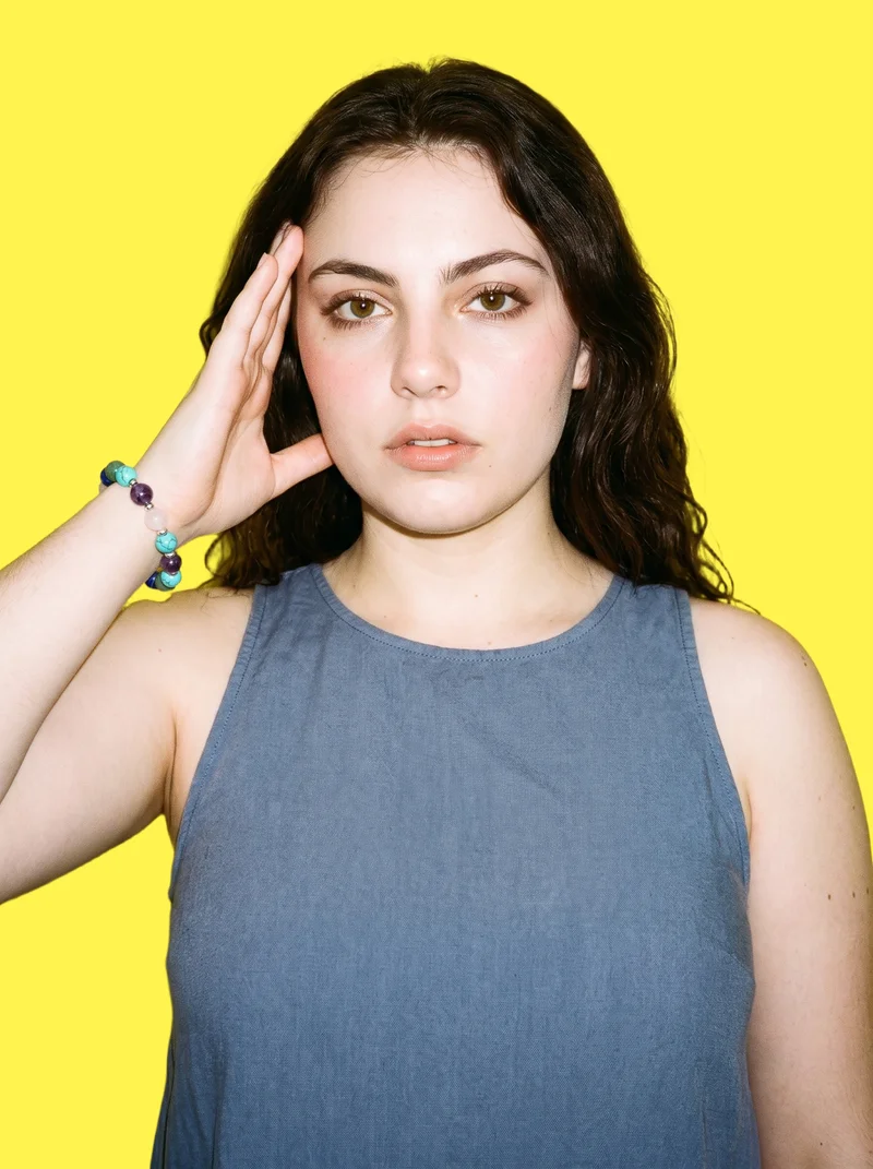A 19-year-old Caucasian woman is shown in an upper-body front view with one hand at her temple and a natural expression, wearing a gemstone beaded bracelet. The image has a flat lemon-yellow backdrop.