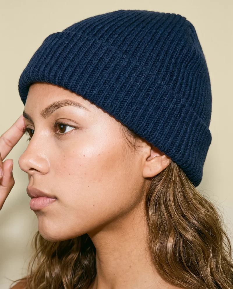 An 18-year-old Hispanic woman is shown in a head-framed side view wearing a navy beanie, with one finger resting at her temple and a serious expression. A wheat-field backdrop is visible behind her.