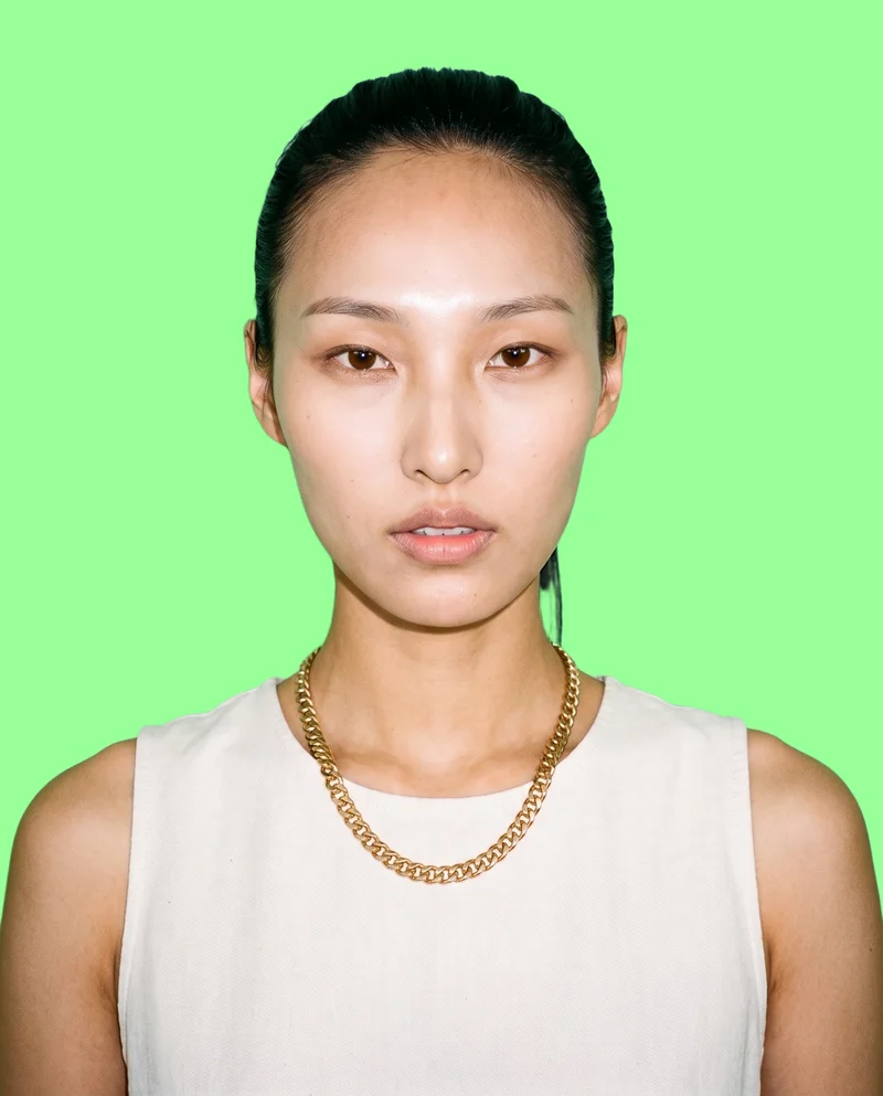 A 27-year-old Korean woman wears a gold chain choker in a front-facing bust view. Her chin is lowered and her expression is natural, with a flat mint green backdrop.