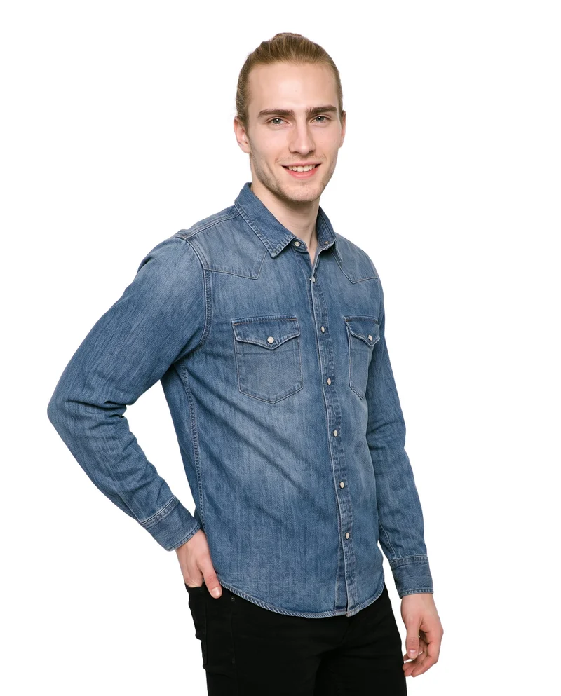 A 20-year-old Caucasian man is shown from the upper body at an angled front view, smiling with one hand on his hip while wearing a mid-wash western denim shirt. The shirt is presented against a pure white backdrop.