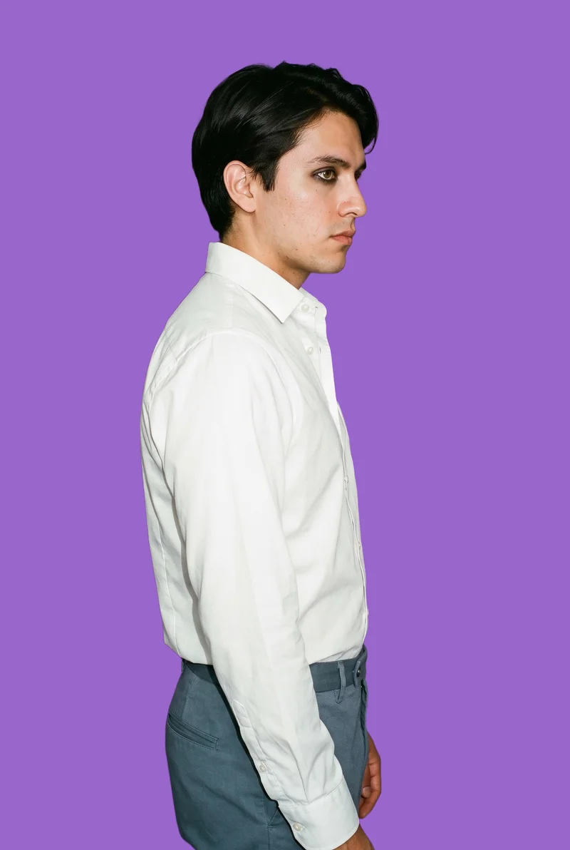 A 29-year-old Hispanic male model wears a white Oxford shirt in an upper-body side view with a shoulder-drop pose and a confident expression. The subject is shown against a flat amethyst backdrop.