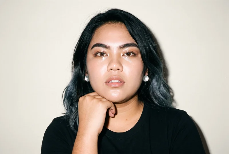 A 23-year-old Southeast Asian woman is shown in a bust-length front view with a neutral expression, wearing women's silver stud earrings. Her wrist is held at her jaw, with an Oyster Pearl studio-wall backdrop.