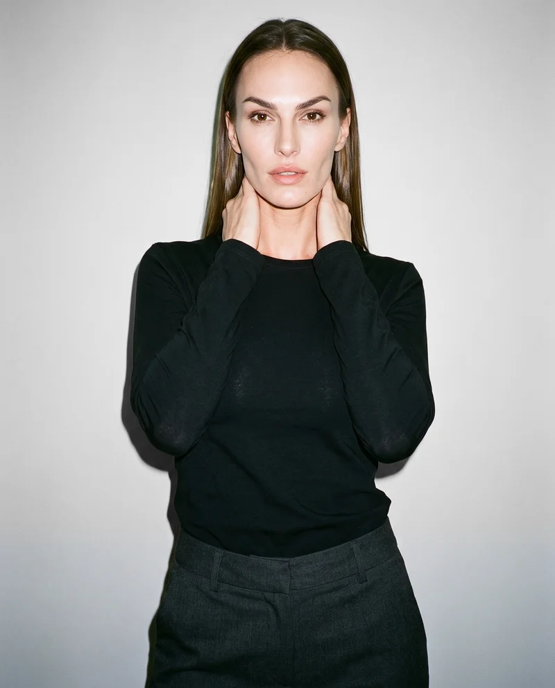 A 35-year-old Caucasian woman is shown from the upper body in a front view wearing a black long-sleeve tee, with her hands on her neck and a confident expression. A pearl grey studio-wall backdrop frames the composition.