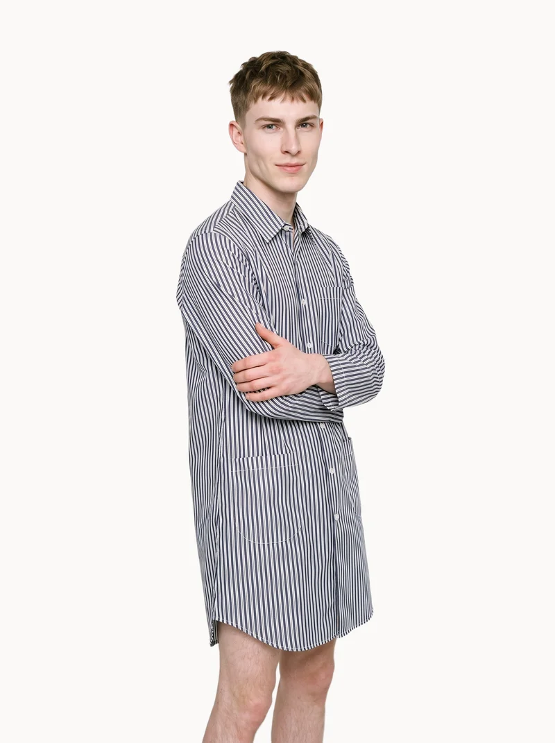 A 25-year-old Caucasian man is shown full body from a front three-quarter angle, wearing a striped nightshirt and holding himself in a self-hug pose. He has a natural facial expression, with a Chantilly Lace flat-color backdrop.