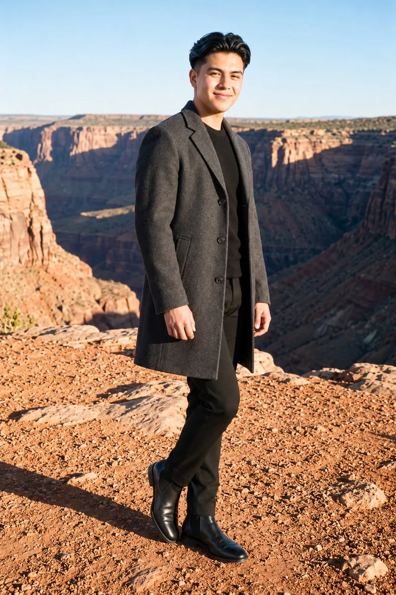 An 18-year-old Hispanic male model wears a men's charcoal overcoat in a full-body, angled-front view with his ankles crossed and a natural expression. The Grand Canyon Rim appears as the backdrop.
