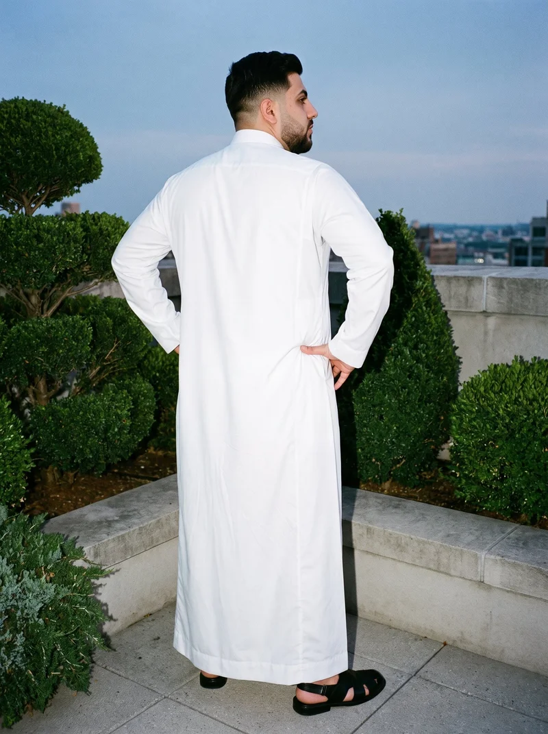 A 26-year-old Arab man wears a white classic thobe in a full-body view, standing with his hands on his hips. The garment is shown from an angled rear view against a rooftop garden backdrop.