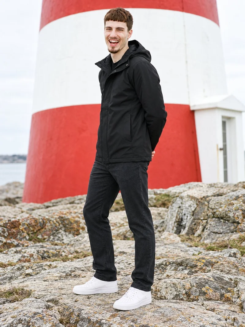 A 25-year-old Caucasian man is shown full body from an angled front view wearing a black hooded raincoat. He stands with his hands behind his back and a laughing expression, against a New England lighthouse backdrop.