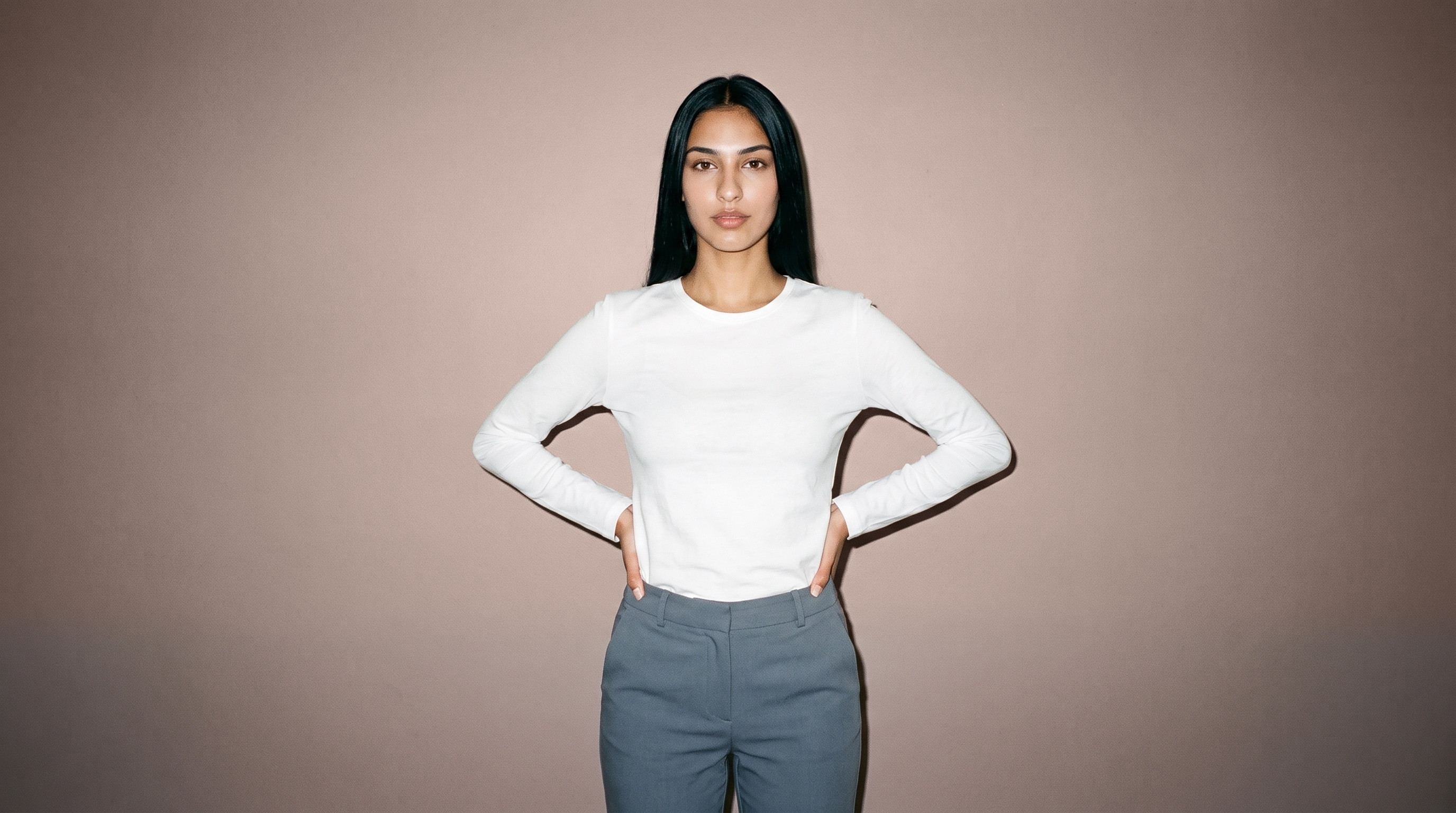 A 19-year-old Turkish woman wears a white long-sleeve tee in a front-facing three-quarter-length view, standing with her hands on her waist and a confident expression. A mushroom-colored studio wall forms the backdrop.
