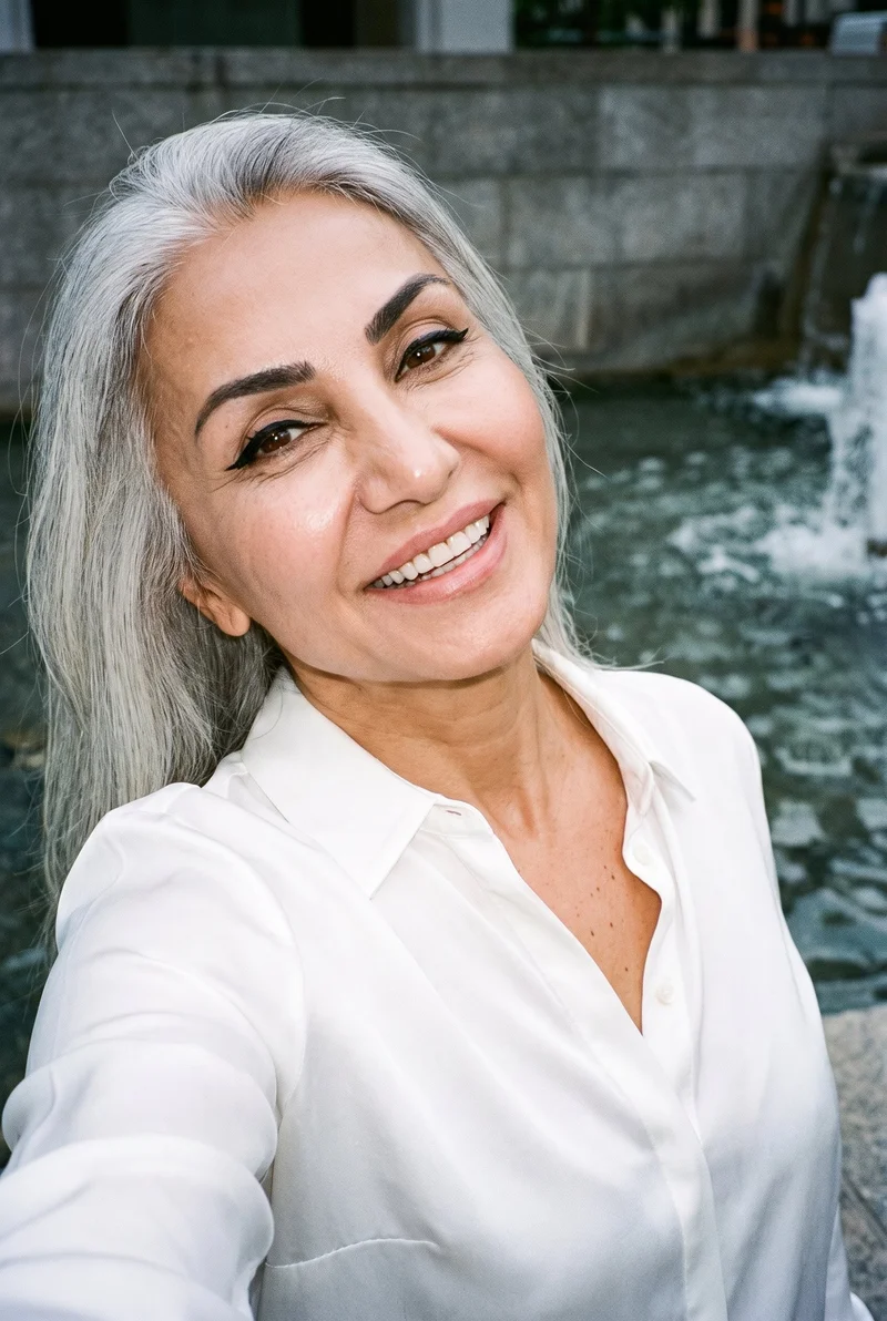 A 58-year-old Turkish woman wears a white silk blouse in a front-facing selfie. Her head is tilted and she is grinning, with a Fountain Plaza backdrop.