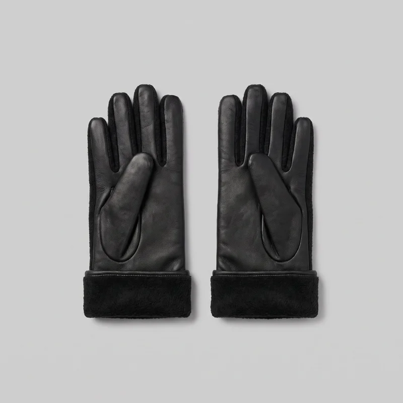 A complete pair of women's black fleece-lined leather gloves is laid flat with the palm side visible. The full-finger design shows separate rounded fingers, inset thumbs, smooth pebbled leather panels across the palms, fingers, and thumbs, fuzzy pile sections along the side edges and between the fingers, and wide plush wrist cuffs with narrow rolled seams. No closures, hardware, logos, prints, embroidery, or embellishments are visible.