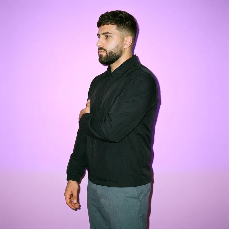 A 22-year-old Caucasian man wears a black windbreaker in a three-quarter-length side view. He is shown holding his arm with a confident expression against a mauve studio-wall backdrop.