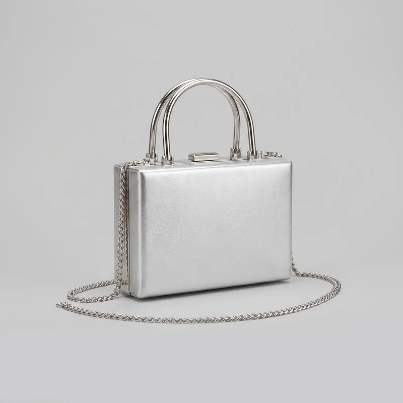 Silver structured box clutch shown in a standalone three-quarter view. The handbag has a rigid rectangular silhouette with softly rounded corners, a flat front panel, boxy side gussets, arched top handles on hinged bracket hardware, and a centered rectangular clasp on the top frame. A chain shoulder strap is attached at the upper sides and draped around the base; edge seams, side hardware, handle mounts, chain links, and a subtly pebbled exterior are visible.