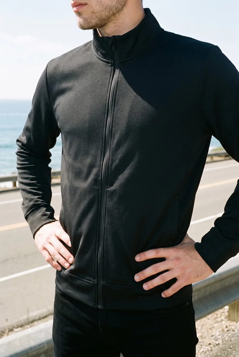 A male model wears a black men's track jacket in a torso-framed, angled front view with his hands on his hips. The Pacific Coast Highway appears as the backdrop.
