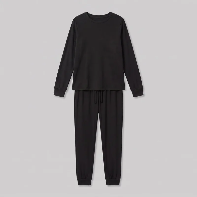 A matching black two-piece loungewear set is laid flat from the front against a backdrop. It includes a full-length ribbed crewneck top with long sleeves, set-in shoulders, a straight hem, and banded cuffs, paired with full-length mid-rise jogger-style pants featuring a visible center-front drawstring, tapered legs, and banded ankle cuffs. Both pieces have an all-over vertical rib texture.