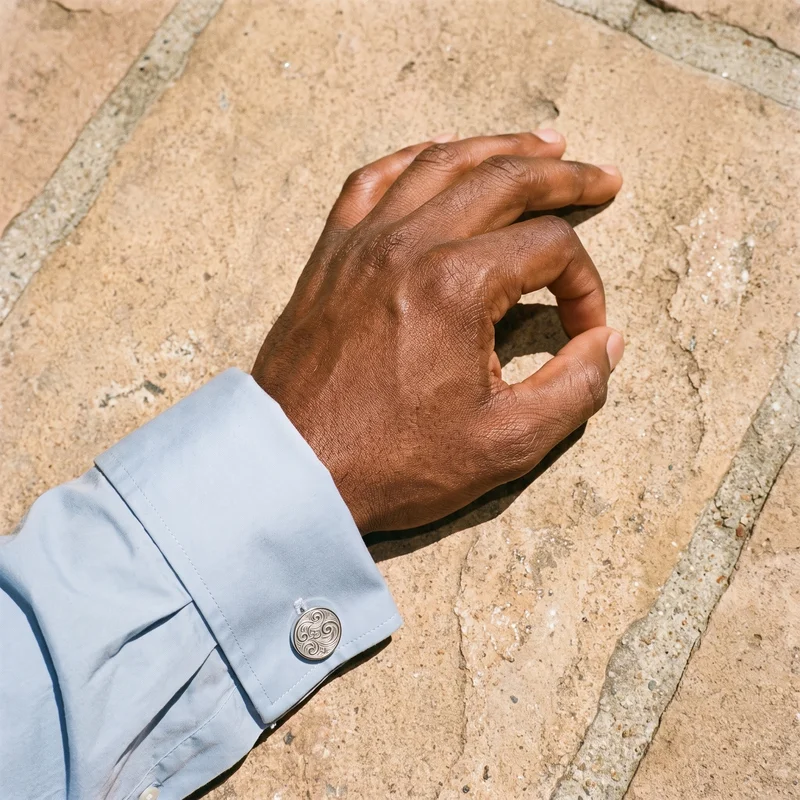 A top view shows a hand and wrist wearing engraved novelty cufflinks, with the thumb touching a finger. The Mission Courtyard Bell Fountain appears as the backdrop.