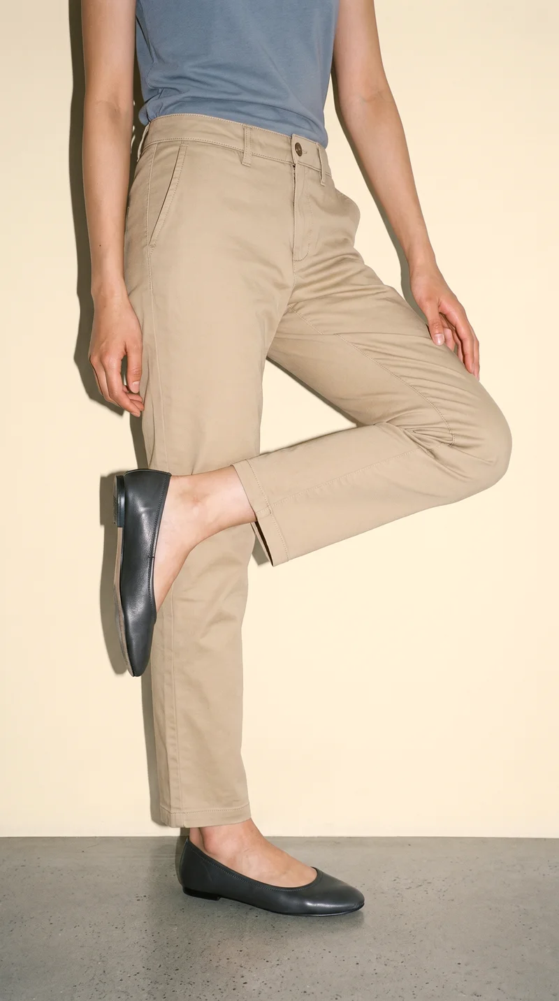 Women's khaki chinos shown on a lower-body model crop from a front three-quarter angle, framed from the feet to the calves. A champagne studio-wall backdrop is visible.
