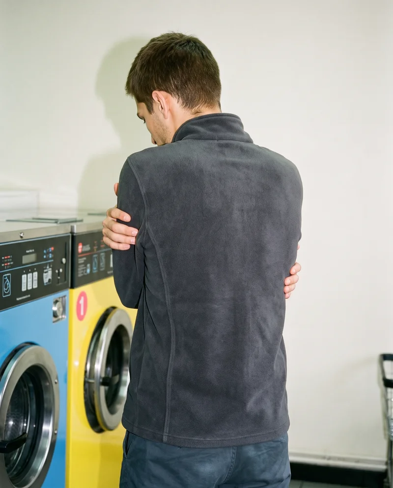 An 18-year-old Caucasian male model wears a black fleece jacket, shown in an upper-body back view with his arms crossed in a self-hug pose. A laundromat serves as the backdrop.