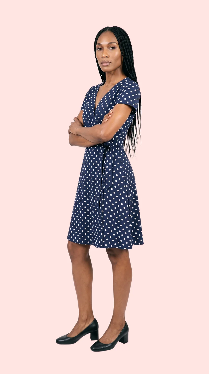 A 42-year-old African woman wears a polka-dot wrap dress in a full-body front three-quarter view. She stands with her arms crossed and a confident expression against a flat Misty Rose backdrop.