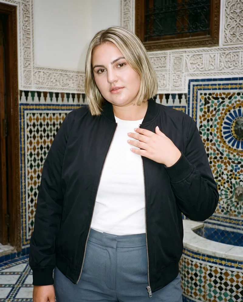 A 21-year-old Caucasian woman wears a black bomber jacket, shown from the upper body at a front three-quarter angle with her fingers at her chest and a natural expression. A Moroccan riad courtyard serves as the backdrop.