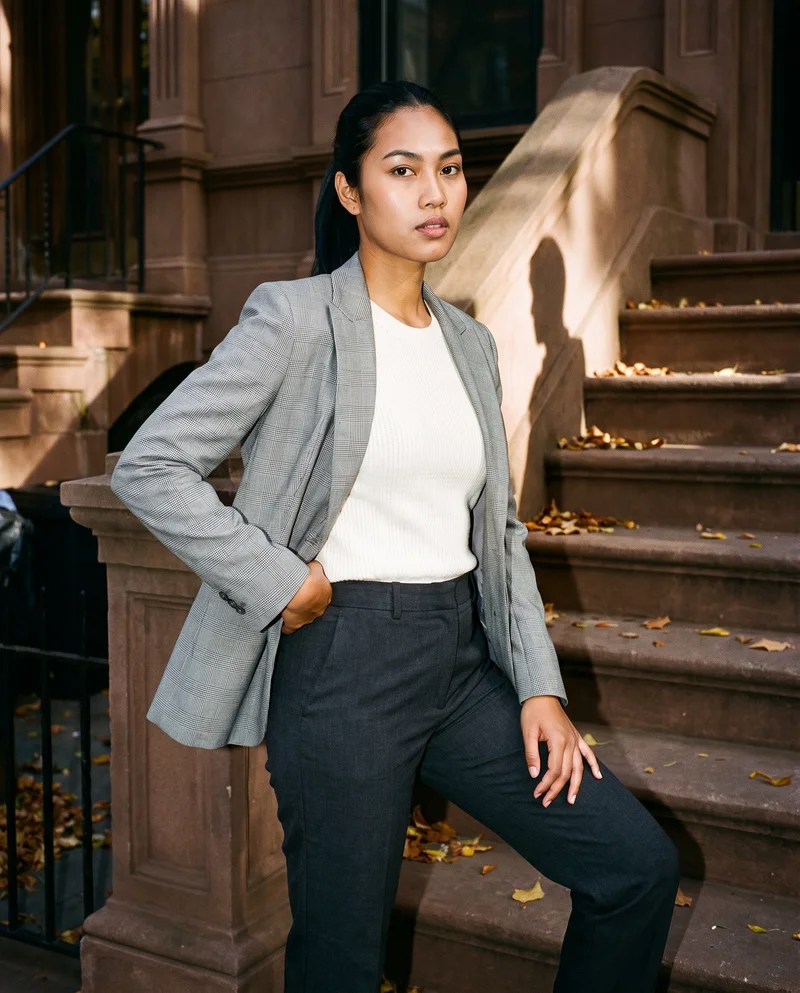 A 19-year-old Southeast Asian woman wears a grey checked blazer in a three-quarter-length view from a front three-quarter angle. She poses with one hip out and a fierce expression, with a brownstone stoop and autumn leaves as the backdrop.