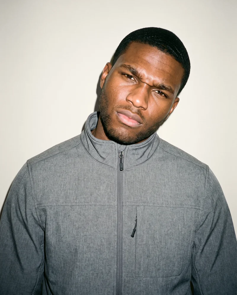 A 22-year-old African man is shown from the front in a bust-length view wearing a men's grey softshell jacket. His head is tilted and he has an intense expression, against an Oyster Pearl studio-wall backdrop.