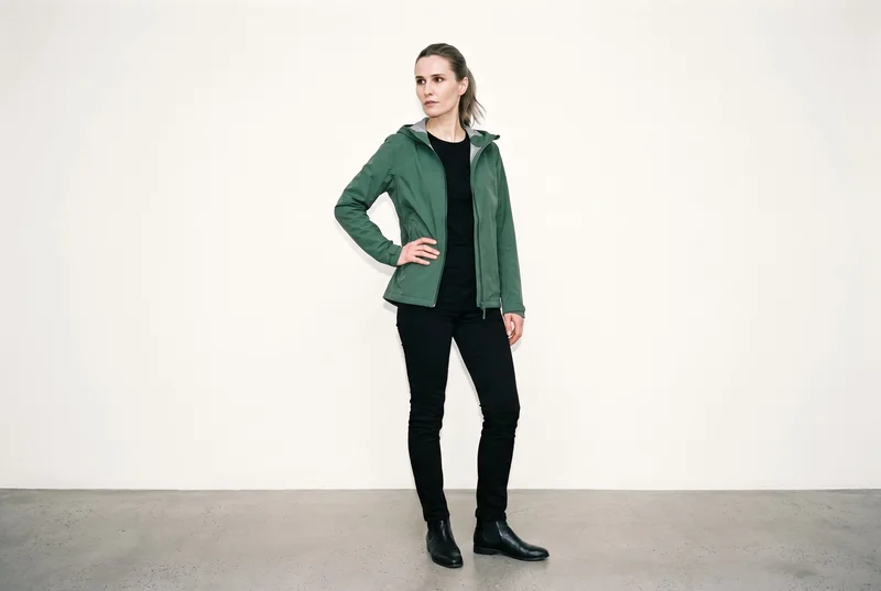 A 39-year-old Slavic woman wears a green waterproof jacket in a full-body, angled-front view. She stands with one hand on her hip and a neutral expression against a soft ivory studio-wall backdrop.